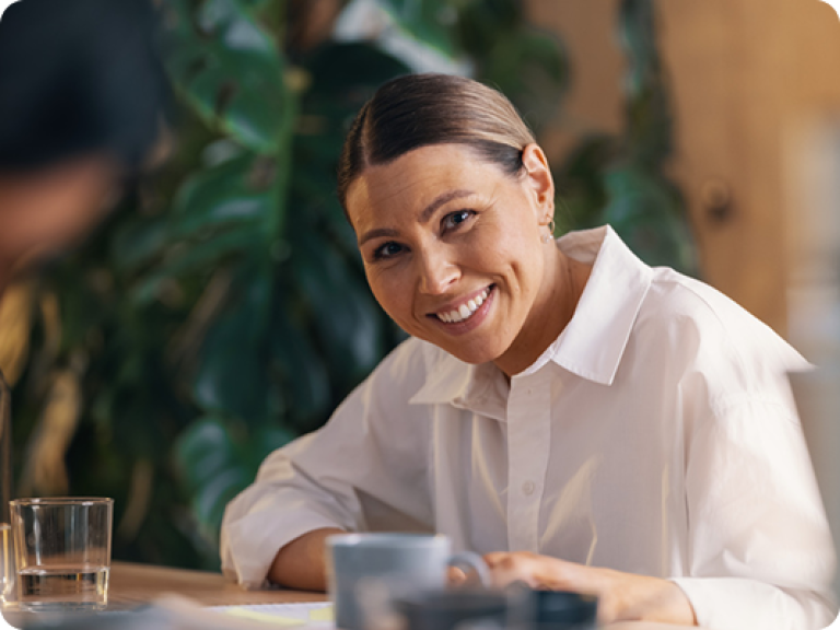 Dame som sitter i et kontorlandskap og jobber, smiler og ser inn i kamera